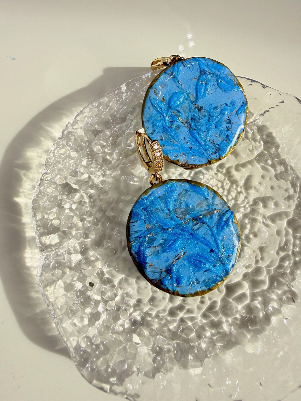 Flora Blue polymer clay statement earrings styled on a textured glass dish in sunlight